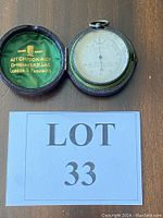 Vintage round pocket barometer inside open protective leather case with green velvet lining and gold branding of Aitchison & Co. visible on the case interior lid.