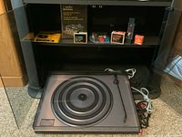Turntable assembly on carpeted floor with shelf backdrop, tonearm and platter visible