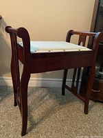 Side view of wood framed bench with slight cabriole legs, scroll top arms, and light beige padded seat.
