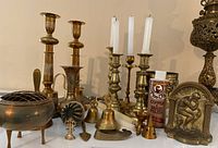 Wide view of the lot showing brass candlesticks with white candles, small brass bells, copper incense burner, trinket box, polish bottle, and brass bookend.