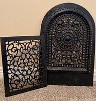 Two cast iron heating grates shown together on floor against wall. One arched with ornate circular design, one smaller rectangular with scrollwork.