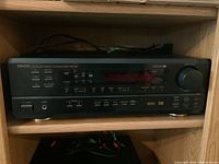 Front view of Denon AVR-1601 receiver set on wooden shelf showing front panel with power button, headphone jack, input buttons, and volume knob.