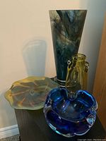 Group photo of four decorative glass items on a table: tall marbled vase, blue glass dish, yellow fluted plate, yellow bottle with handles.