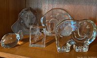 Shelf display photo showing all five clear glass figurines – lion, fish, geese block, elephant, and book-shaped piece.