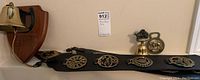 Wide shot showing brass hand bell mounted on wooden plaque, brass bottle opener, small brass bell, and two leather straps with multiple brass horse brasses attached.