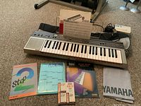 Full view of Yamaha PC-1000 keyboard on floor with integrated music stand, sheet music cards, instruction manuals, music books (Step 2, Yamaha Play Now), and Yamaha branded soft carry case visible around keyboard.