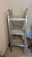 Folded aluminum step ladder shown upright in storage position, displaying multiple steps and aluminum frame.