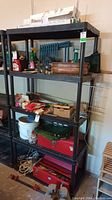 Front view of black plastic shelving unit with five shelves, empty, standing in a garage environment next to a wall with various stored items nearby but not on shelves.