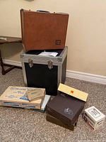 View of brown leather carrying case with Kodak logo atop a metal storage case with various boxes and slide accessory packets on the floor.