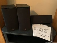 Photo of two black Klipsch SB-1 bookshelf speakers and one horizontal SC-5 centre speaker on a shelf with manuals included.