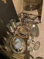 Photo showing multiple silver plated serving trays, goblets, candle holders with glass chimneys, monogrammed sterling silver napkin rings, and salt cellars arranged on floor near corner.