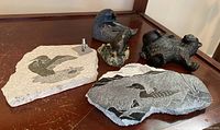 Four stone and coal carvings on wooden surface: a bronze bear sculpture on stone base, two soapstone loon carvings on stone slabs, and a reclining bear carved from coal.