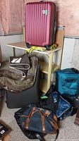 Collection of suitcases and bags displayed on and around a small table. Includes large red hard-shell suitcase, brown and black duffle bags, blue soft suitcase, and yellow luggage straps.