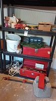Three-tier black shelving unit with assorted items including red toolboxes, white bucket, gloves, and electrical cords.