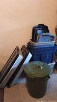 Side view of stacked Rubbermaid storage totes in navy blue and gray with lids placed on top and to the side, plus green Rubbermaid garbage can in front.