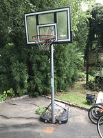 Photo shows a single adjustable-height basketball hoop outdoors on a driveway/paved surface with greenery and bicycles around. The backboard is large and rectangular with a smaller inner rectangle, red rim, and attached net. The pole is metallic with some rust and connects to a base.