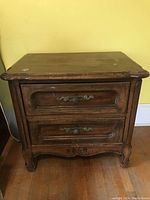 Front view showing two drawers with carved panels and ornate metal handles. Top surface has minor scratches.