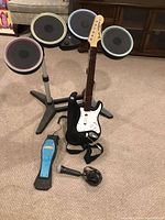 Photo showing Rock Band PS2 game set with guitar controller, drum set, foot pedal, and microphone on carpeted floor.