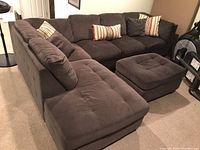 Photo showing a U-shaped sectional sofa in gunmetal grey fabric with tufted cushions and matching ottoman, accompanied by three coordinating pillows.