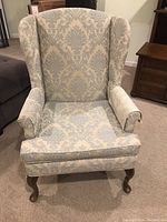 Front view of the jacquard upholstered wing chair showing the damask fabric pattern, wing arms, and ball-foot wooden legs with visible wear on armrests and legs.