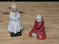 Two Royal Doulton porcelain figurines of small children: one kneeling in a white nightgown praying, one sitting wrapped in a red floral blanket.