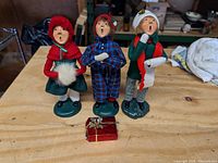 Photo showing three Byer's Choice caroler figurines standing on a wooden surface including a girl in a red cloak, a boy in blue plaid pajamas, and another boy in red and green vest holding a songbook.