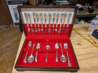 Open mahogany chest lined with red velvet holding selected flatware pieces, showing detailed handle pattern.