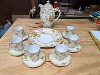 Full set arranged on wooden table showing chocolate pot, plate, and 4 cups with saucers, featuring ivory background and floral pattern.