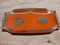 Top-down photo of a rectangular orange Georges Briard glass tray with gold decorative elements and teak handles on sides, placed on wooden surface.