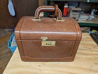 Front view of the brown leather makeup and jewelry case showing the brass combination lock and handle on top.