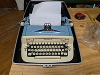 Full view of blue Royal portable typewriter with white paper inserted, housed in black carrying case on wooden floor.