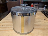Front view of ice bucket showing the textured metal surface with vertical gold accent strip and the clear lid with decorative knob.