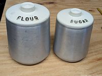Set of two mid-century kitchen cannisters labeled FLOUR and SUGAR with aluminum bodies and white lids.