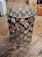 Full view of cylindrical side table showing glass top and mirrored circular accents on metal frame, on wood floor background.