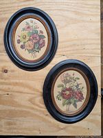 Two black oval picture frames with floral prints laid flat on wood surface