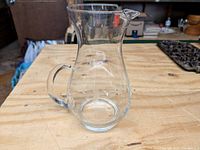 Front side view of a transparent glass pitcher with handle and spout sitting on a wooden surface.