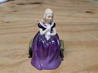 Front view of Royal Doulton porcelain figurine of a girl in a purple dress holding a white cat while seated on a small green cushioned bench.