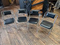 Four Samsonite folding chairs arranged in semi-circle showing black vinyl cushions and chrome frames on wood floor.