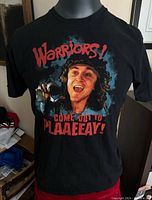 Front view of the black T-shirt showing a vibrant graphic print featuring a character from The Warriors movie and text 'Warriors! Come Out to Plaaeeay!'