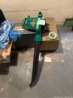 Green handheld Barracuda blower/mulcher/weed eater device with black blower tube lying on a box in a basement.