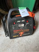 Photo of black MotoMaster Eliminator 500A Booster Pack unit with cables attached sitting on floor in garage setting.