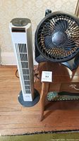 Photo showing the full view of the two fans side by side: Honeywell black desk fan on wooden table and Hampton Bay white tower fan on floor.