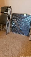 Photo showing four stacked metal frame folding chairs with black padded seats and backs next to a folded rectangular table wrapped in plastic sitting vertically against the wall.