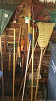 Various garden rakes, brooms, and hand tools leaning against a wooden wall in storage.
