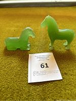 Front view of two green jade horse figurines, one standing and one lying down on a yellow surface with auction tag.
