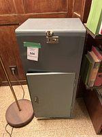 Full view of the metal cabinet showing front door with lock and top lid with latch, dark gray finish, placed on a tiled floor next to bookshelves and a floor lamp.