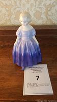 Front angled view of Royal Doulton porcelain figurine seated girl in purple dress on a wooden surface.