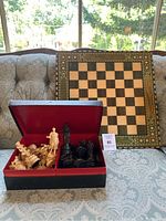 Overall view of chess board leaning on couch with red lined box containing black and white chess pieces in front.