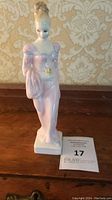 Front view of the Daphne figurine with pink gown and yellow flower detail on waist, standing on wood surface with wallpaper background.