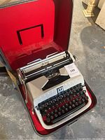 Vintage off-white manual typewriter in open hard case with red fabric lining, showing overall condition and carry case.